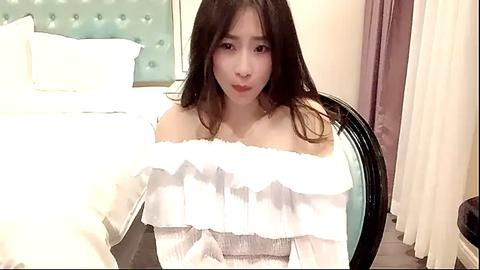 Media: A video of an East Asian woman with long, dark hair, wearing an off-shoulder white dress, sitting on a chair in a bedroom with a tufted headboard, pale blue curtains, and a black-framed mirror.