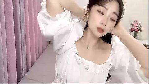 Media: Video of a young Asian woman with light skin, long black hair, and delicate facial features, wearing a white, ruffled blouse, posing in a softly lit bedroom with pink curtains and a vase of flowers on a nightstand.