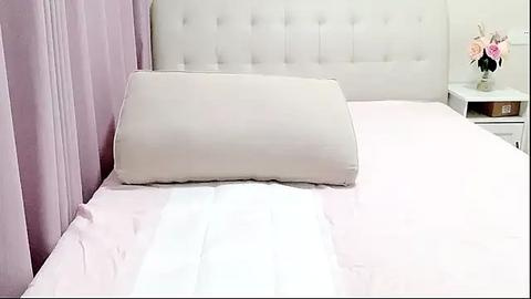 Media: Video of a neatly made bed with a white tufted headboard, light pink bedding, and a white pillow, set against a lavender curtain backdrop.