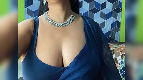 Media: Video of a woman with medium skin tone, large breasts, wearing a deep blue sleeveless top with a plunging neckline. She has a turquoise necklace. Background features a blue and white geometric pattern.