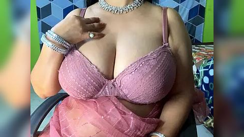 Media: Video of a voluptuous woman with medium skin tone wearing a pink lace bra and sheer pink skirt, adorned with silver jewelry, seated in a colorful chair.