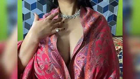 Media: A video of a woman with medium skin tone, wearing a low-cut, vibrant pink paisley-patterned scarf over a revealing neckline, adorned with a diamond necklace. She has a manicured hand touching her chest. Background features a geometric, blue-and-white tiled wall and a green wall.