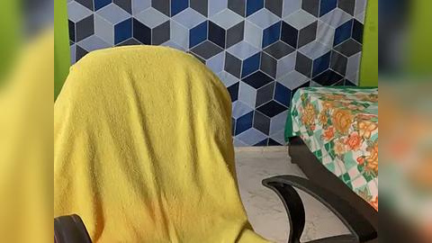 Media: A video of a chair with a yellow towel draped over it, next to a bed covered with a floral-patterned green and orange bedspread, in a room with a blue and black hexagonal-patterned wall.