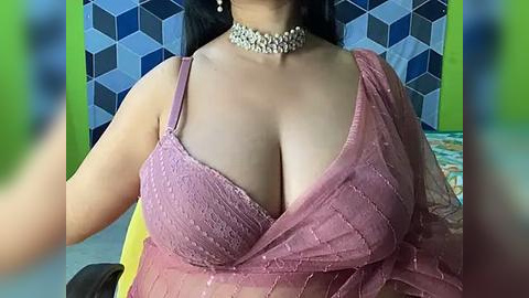 Media: A video of a woman with medium skin tone, wearing a pink, semi-sheer lace bra and a silver choker. She has long, dark hair and is standing against a geometric blue and black backdrop.