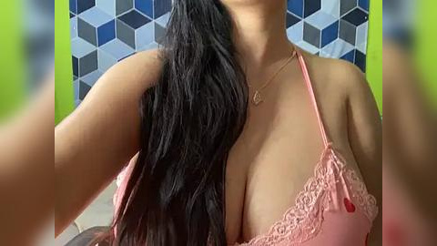 Media: Video of a woman with long black hair, wearing a pink lace bra with thin straps, her cleavage partially visible. Background features a geometric patterned wall.