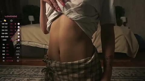 Media: Video of a woman lifting her white shirt, revealing her midriff and wearing plaid pajama bottoms. Background shows a dimly lit bedroom with a bed and potted plants.