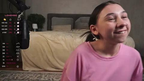 Media: Video of a smiling young girl in a pink shirt, standing near a gray bed with beige bedding, in a dimly lit bedroom.