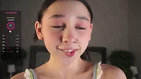 Media: Video of a young woman with light skin, brown hair tied back, smiling with closed eyes, wearing a white tank top, and a septum ring. Background is blurred, with a digital interface overlay showing \"LOVENSE\" and \"Real-time feedback.\