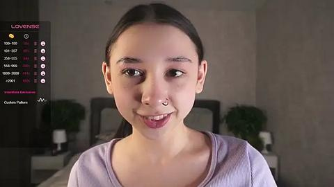 Media: Video of a young Asian woman with long black hair, light skin, wearing a light purple shirt, smiling, in a modern bedroom with a white bed and green plants.