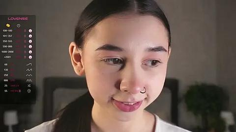 Media: Video of a young Asian woman with straight, dark hair, light skin, and a nose ring. She has a neutral expression. Background shows a blurred, dimly lit room with a green plant.