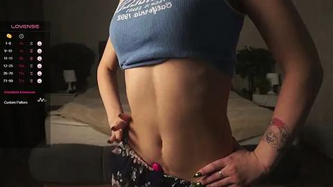 Media: Video of a woman in a blue ribbed crop top, revealing a flat stomach and tattooed left wrist. Background shows a dimly lit bedroom with a bed and potted plant.