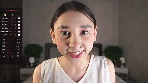 Media: Video of a young Asian woman with straight black hair, fair skin, and a silver nose ring, wearing a white tank top. Background shows a dimly lit bedroom with a bed, nightstand, and potted plant.