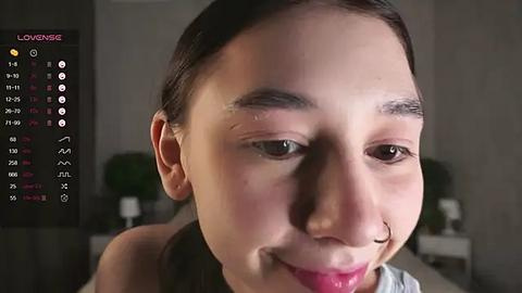 Media: A close-up video of a smiling woman with light skin, dark brown hair, and a nose ring, wearing a light-colored top, in a dimly lit room with a calendar app on the left side.