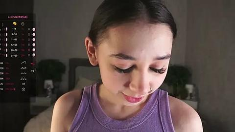 Media: Video of a young Asian woman with straight black hair, wearing a sleeveless purple top, in a dimly-lit bedroom with a bed and green potted plants in the background.