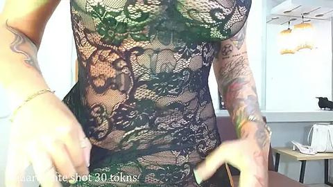 Media: Video of a tattooed person wearing a sheer, black lace bodysuit with floral patterns, showcasing their muscular torso and arms, in a modern, brightly-lit room with a white wall and wooden table.