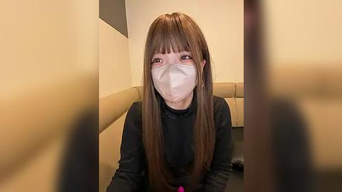 Media: Video of an Asian woman with long brown hair and bangs, wearing a beige mask, black turtleneck, and sitting on a beige cushioned seat. The background features beige walls and a blurred figure in the foreground.