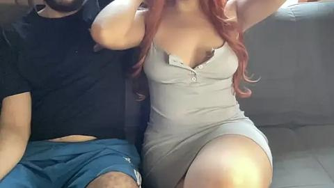 Media: Video of a curvy, light-skinned woman with long, wavy red hair wearing a tight, low-cut grey dress with a zipper, sitting on a grey couch. Her breasts are prominently exposed. Next to her, a dark-skinned man in a black shirt and blue shorts sits, his face partially visible.