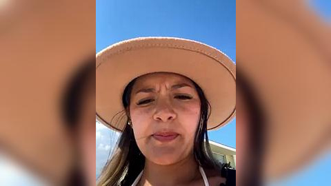 Media: Video of a young Asian woman with long brown hair, wearing a beige sun hat, squinting against bright sunlight.