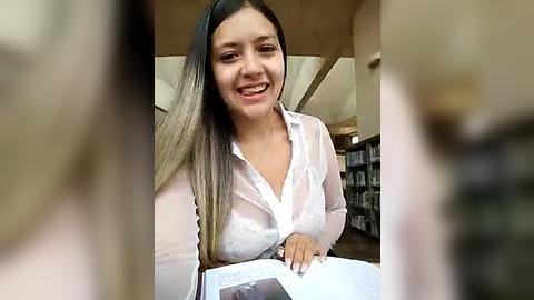 Media: Video of a smiling Latina woman with long, straight black hair and fair skin, wearing a white blouse, seated at a desk in a library, holding a book.