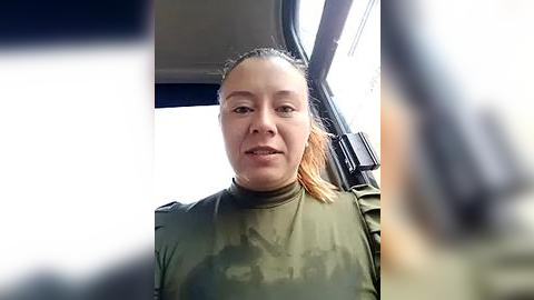 Media: Video of a young woman with light skin and long, light brown hair tied back, wearing a green camo t-shirt, seated in a car. Out-of-focus background features a blurred, white vehicle.