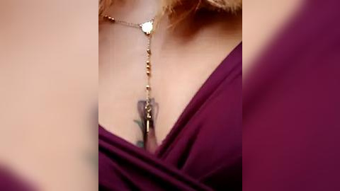 Media: Video of a close-up of a woman's chest, wearing a deep maroon, off-the-shoulder dress with a delicate gold necklace featuring a heart pendant and a feather dangle. The background is blurred.