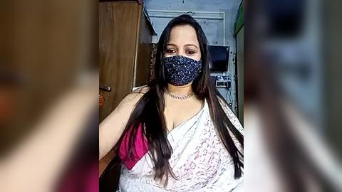 Media: A video of a South Asian woman with long dark hair, wearing a blue floral saree and a blue mask, standing in a dimly lit room with wooden furniture and a TV in the background.