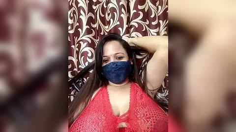 Media: Video of a Latina woman with medium skin tone, long black hair, and a blue mask, wearing a red lace bra, sitting against a patterned brown curtain.