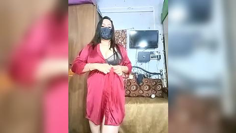 Media: Video of a woman in a red silk robe with a black mask, standing in a dimly lit room with a bed, TV, and a patterned pillow.