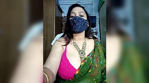 Media: Video of a woman with medium skin tone, wearing a pink lace bra, green sari with floral patterns, blue mask, and gold necklace, standing indoors with blurred background.