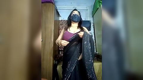 Media: A video shows a woman with long dark hair, wearing a black mask and a purple top, sitting on a bed, holding a black saree. The background reveals a cluttered room with wooden furniture and a TV.