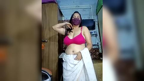 Media: Video of a South Asian woman with medium skin tone, wearing a pink bra and face mask, standing in a cluttered room with a TV and shelves, holding a white cloth.