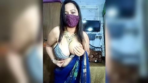 Media: A video of a woman with long dark hair, wearing a blue and gold traditional outfit with a purple face mask, adjusting her cleavage in a dimly lit room with a TV in the background.