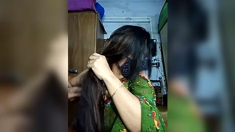 Media: A video captures a woman with medium-length black hair, wearing a green floral shirt and a black face mask, combing her hair in a dimly lit room with a wooden cabinet and a green door.