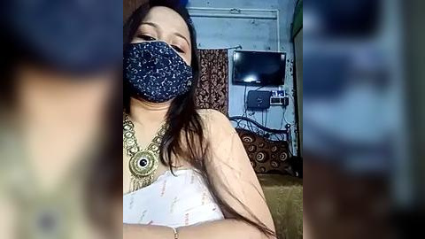 Media: Video of a woman with long dark hair, wearing a blue face mask and ornate gold necklace, sitting in a dimly lit room with a TV and patterned curtains.