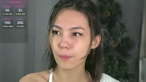 Media: A video of a young Asian woman with straight, shoulder-length dark hair and light skin. She wears a white top. Background shows a blurred plant.