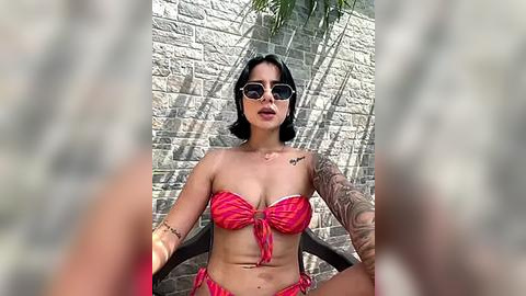 Media: Video of a dark-haired woman with sunglasses, medium breasts, and a tattooed arm, wearing a red and white striped bikini, sitting on a wooden chair in front of a stone wall.