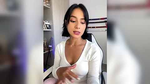 Media: Video of a young woman with medium-dark skin, straight black hair, and a slim build, wearing a white V-neck top, sitting at a desk in a modern office with shelves, a chair, and a whiteboard in the background.