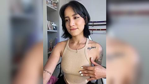 Media: Video of a young woman with short black hair, fair skin, and tattoos on her arms. She's wearing a beige tank top and sitting in a room with shelves and a bed in the background.