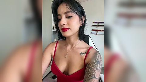 Media: Video of a young woman with medium brown skin and long black hair, wearing a red spaghetti-strap top, showcasing a tattoo sleeve on her left arm, and red lipstick, seated indoors with blurred background.