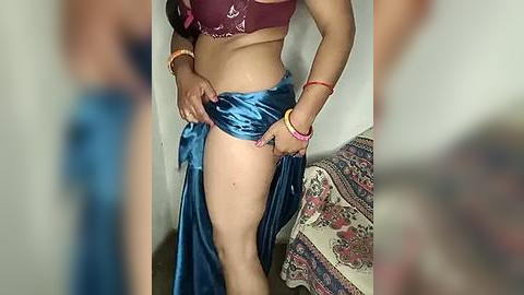 Media: Video of a woman with medium skin tone, wearing a purple bra and blue satin skirt, lifting the skirt to reveal her bare thighs. She's in a room with a colorful patterned blanket and a yellow wall.