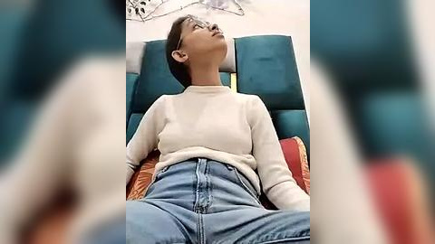 Media: Video of an Asian woman with short dark hair, wearing glasses, a white turtleneck, and high-waisted blue jeans, lying on a teal sofa with red and orange pillows, gazing upward in a relaxed, contemplative pose.