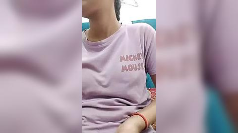 Media: Video of a young woman with medium skin tone, wearing a pastel pink shirt with \"I AM NOT MOODY\" text, seated in a teal chair, blurry background.