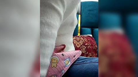 Media: Video of a person's legs in white socks, wearing a pink blanket with cartoon characters, sitting on a blue seat with a red, gold-patterned pillow in a bus.