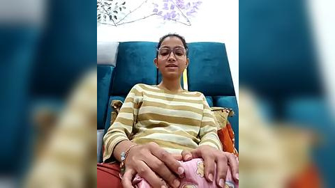 Media: Video of a South Asian woman with medium brown skin, in a striped shirt, sitting on a teal sofa with her hands folded, surrounded by blurred blue and yellow objects in a modern, indoor setting.