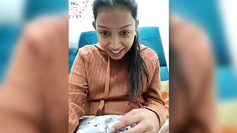 Media: Video of a young Indian woman with medium skin tone, straight black hair, and a bindi, wearing a rust-colored sweater, sitting on a blue couch, holding a phone, in a casual indoor setting.