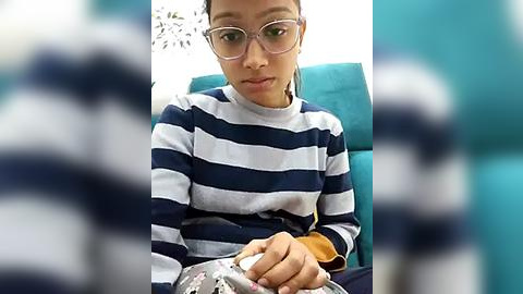 Media: Video of a young woman with short hair, wearing glasses and a navy-blue and white striped sweater, holding a cat. Background is out of focus.