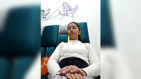 Media: A video of a woman with medium brown skin and black hair, wearing a white sweater and black skirt, sleeping in a teal armchair. Purple flower arrangement and orange pillow in the background.