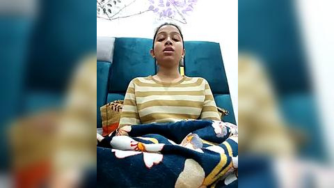 Media: Video of a woman with dark hair and light skin, wearing a striped sweater, lying on a teal couch with a blue blanket featuring floral patterns. She appears relaxed with eyes closed.