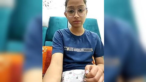 Media: Video of a young girl with light brown skin, wearing glasses and a blue T-shirt with white text, sitting on a teal cushioned chair in a brightly lit room.