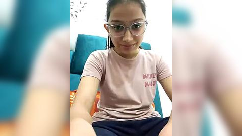 Media: Video of a young Asian girl with glasses, wearing a pink t-shirt and blue shorts, sitting on a teal chair, smiling. Background shows a white wall and blurred people.